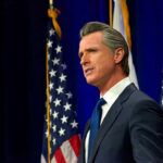 GUILTY: Newsom’s Top Gatekeeper Admits Federal Fraud… Man speaking at podium with American flags behind him.