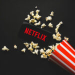 Netflix SURVEILLANCE Exposed — Spying on Children… Spilled popcorn, phone displaying Netflix logo, striped popcorn container.