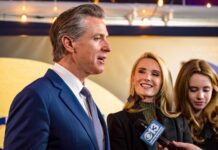 Newsom’s Airport THREAT — Travelers Caught In Crossfire… A man in a suit speaking at an event with two women nearby, one smiling and the other holding a microphone