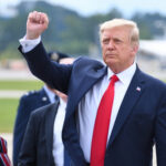 Assassin Hunted Trump’s Inner Circle A political figure raising a fist in a gesture of confidence during an outdoor event