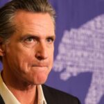 Trump DOJ Heat Hits Newsom’s Home A serious-looking political figure at a press event with a bear graphic in the background